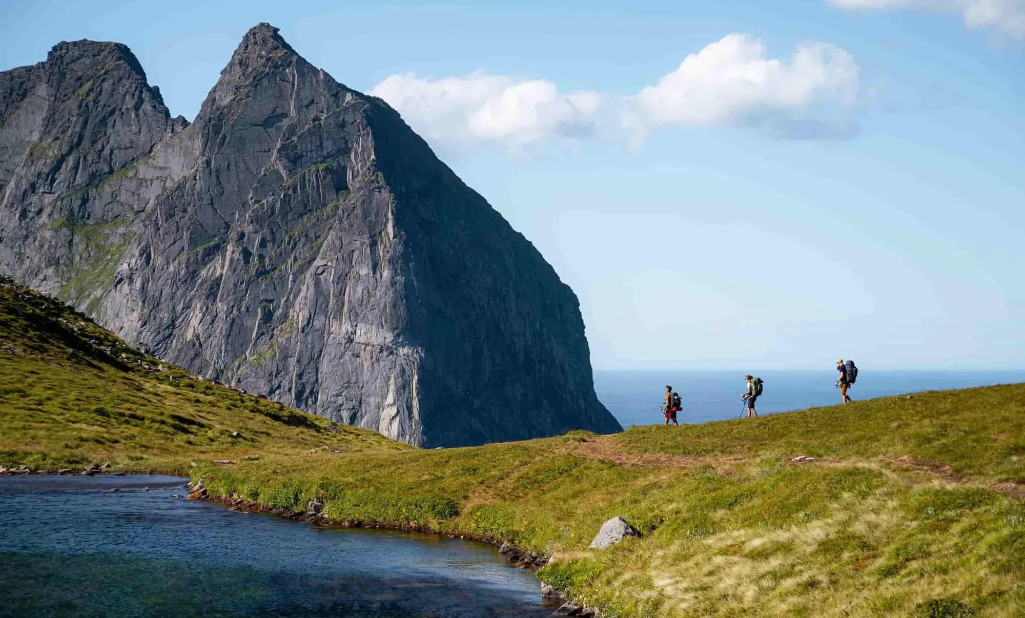 Three hikers crossing a grassy ridge below a granite peak in Lofoten
